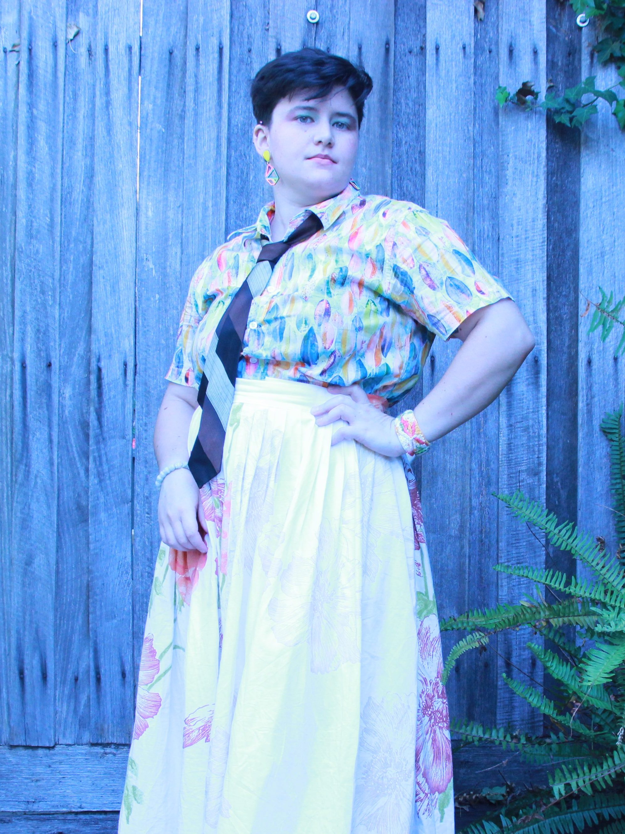 full length photo of a white woman in confident pose with short brown hair, wearing a long skirts button up t-shirt, and a tie, and colorful make-up.
